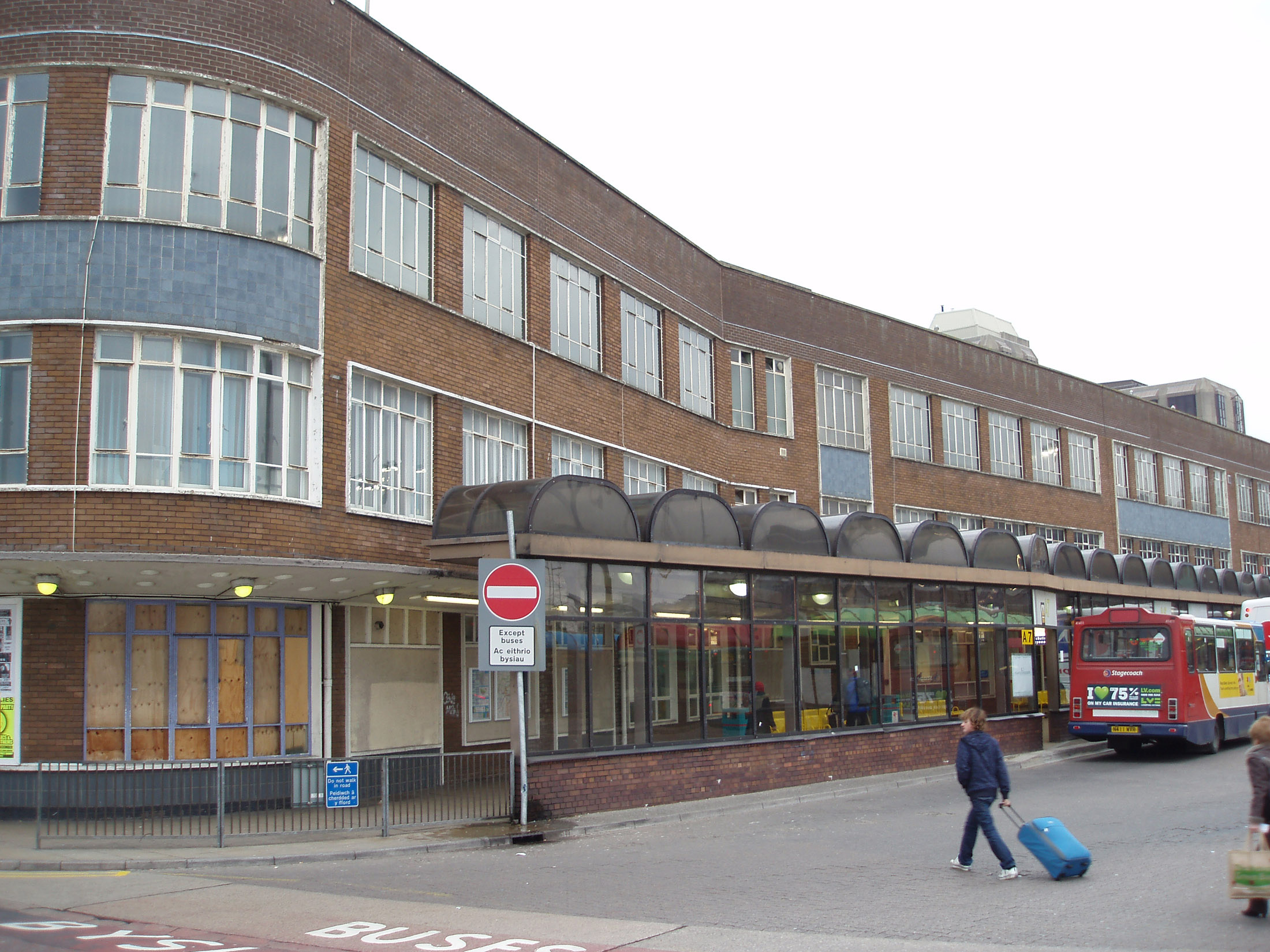 an image of View of the Central Bus Station, Cardiff, Wales with a row of shelters along the exterior of the depot and a woman walking past with her suitcase as a bus loads passengers in the background