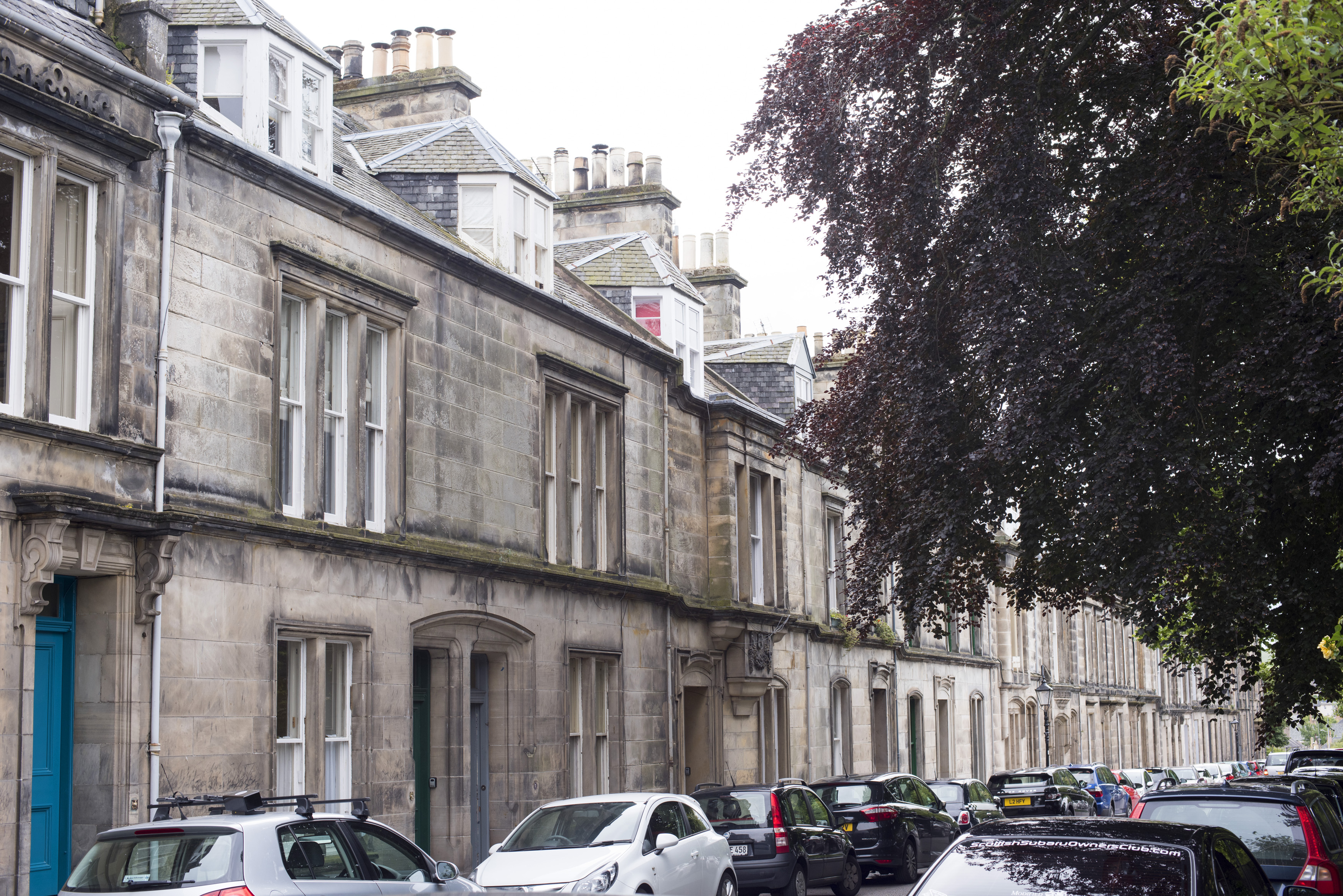 an image of View down street with dark leafed tree hanging over cars in Saint Andrews, Scotland