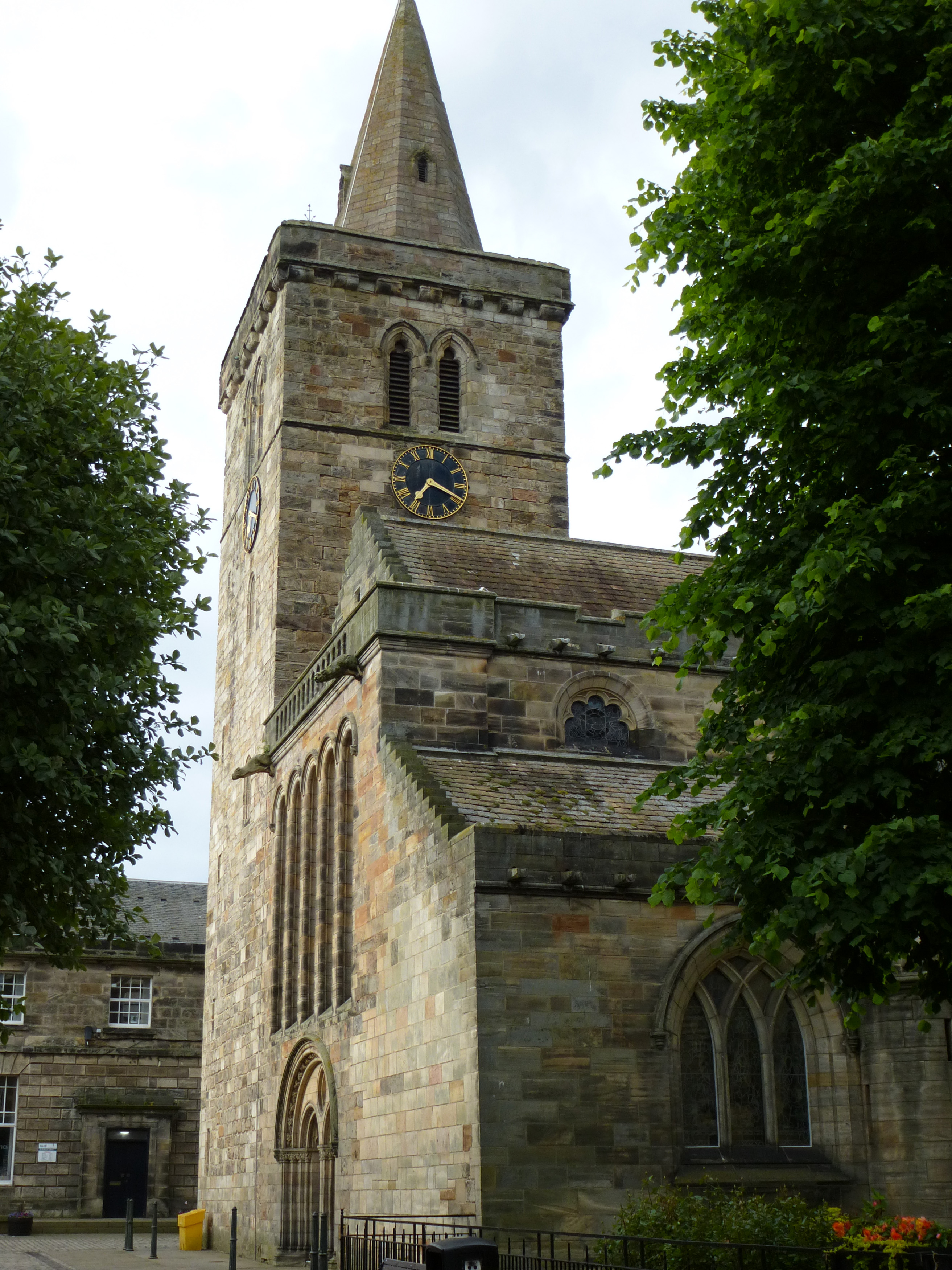 an image of Street level view of tall rectangular clock and bell tower on Holy Trinity Church framed by trees in Saint Andrews, Scotland