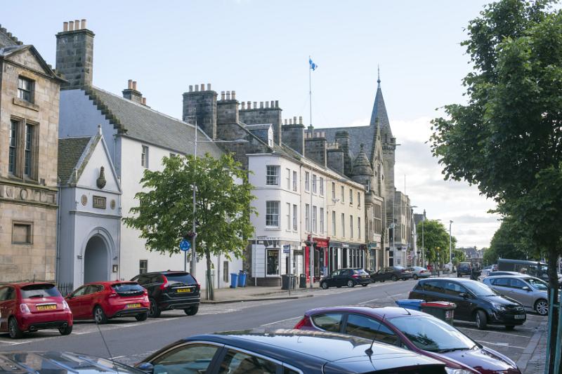 Street scene in St Andrews, Scotland with rows of parked cars in front of historic buildings on a quiet sunny day