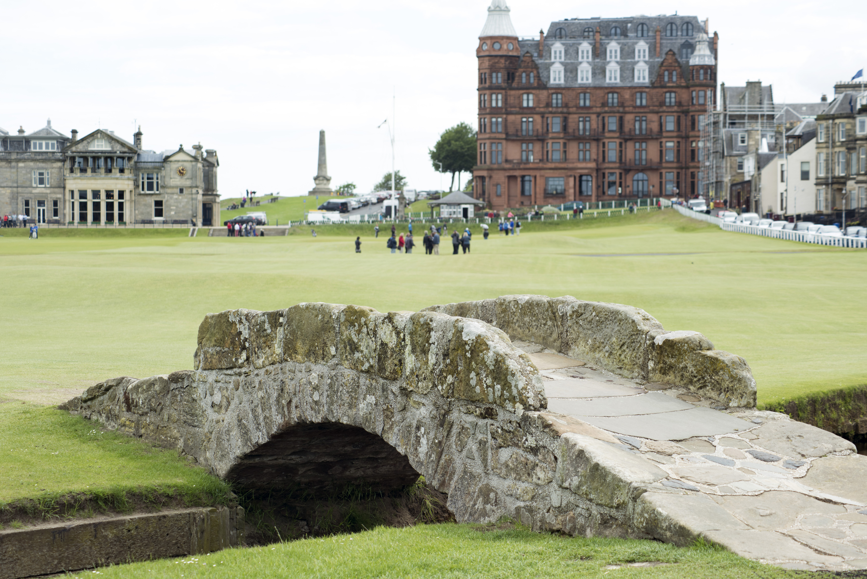 an image of Swilken Bridge, St Andrews golf course, Scotland with a view of the town behind and a group of people on the fairway