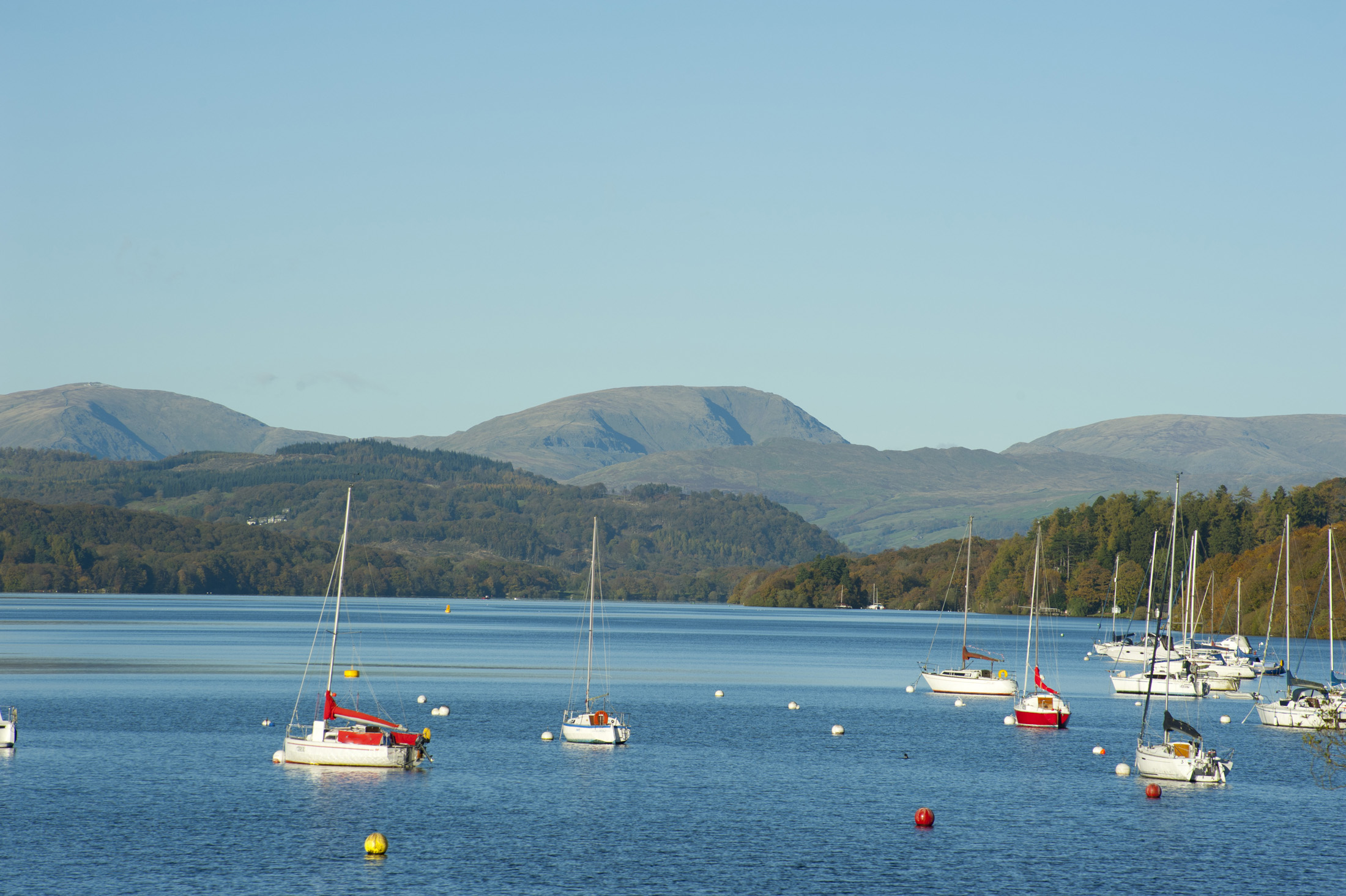 an image of Pleasure yachts moored on the calm water of Lake Windermere in the Lake District in Cumbria against a mountain backdrop