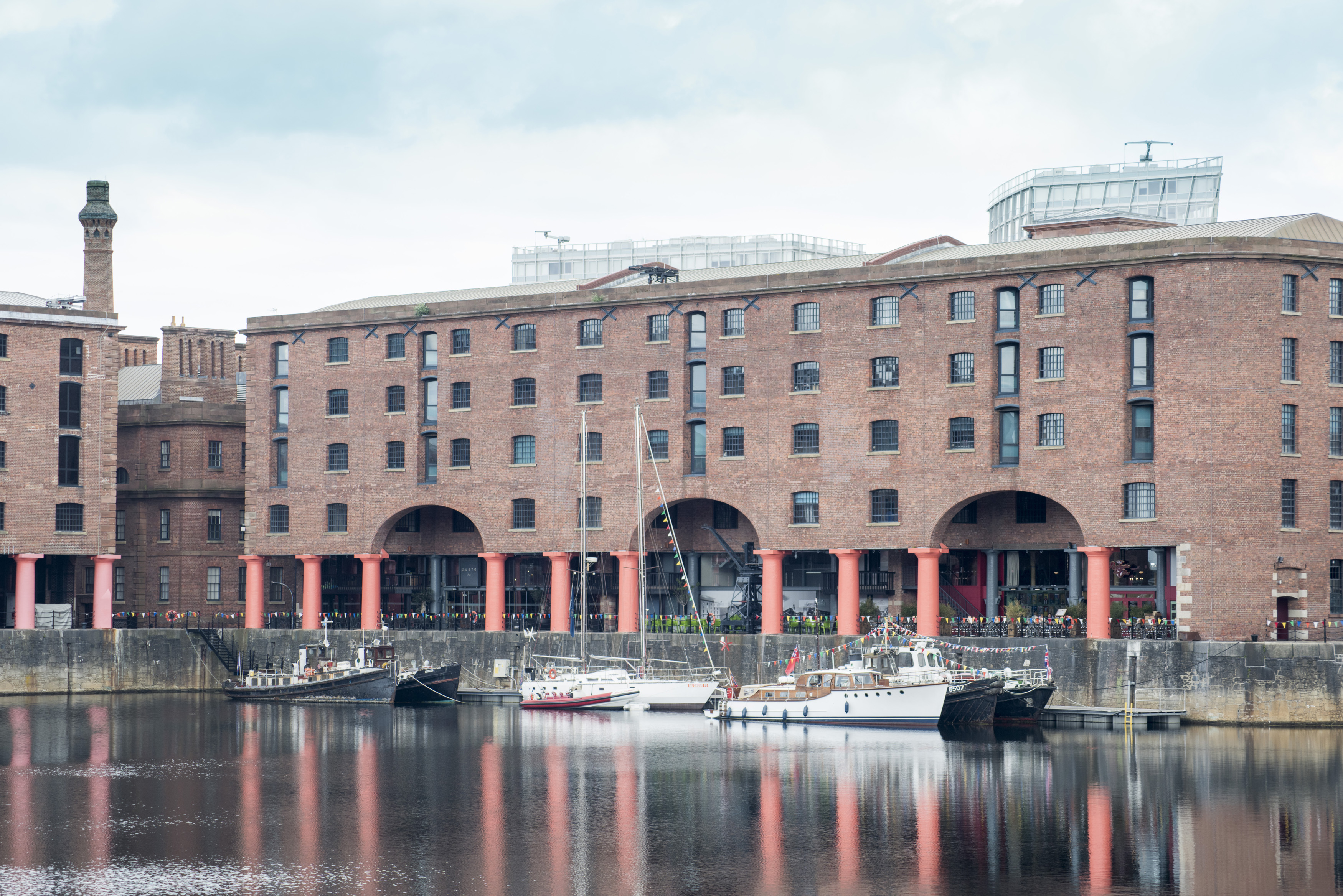 an image of View from across the bay of three small boats docked at Liverpool Albert Dock in front of brick building with red columns