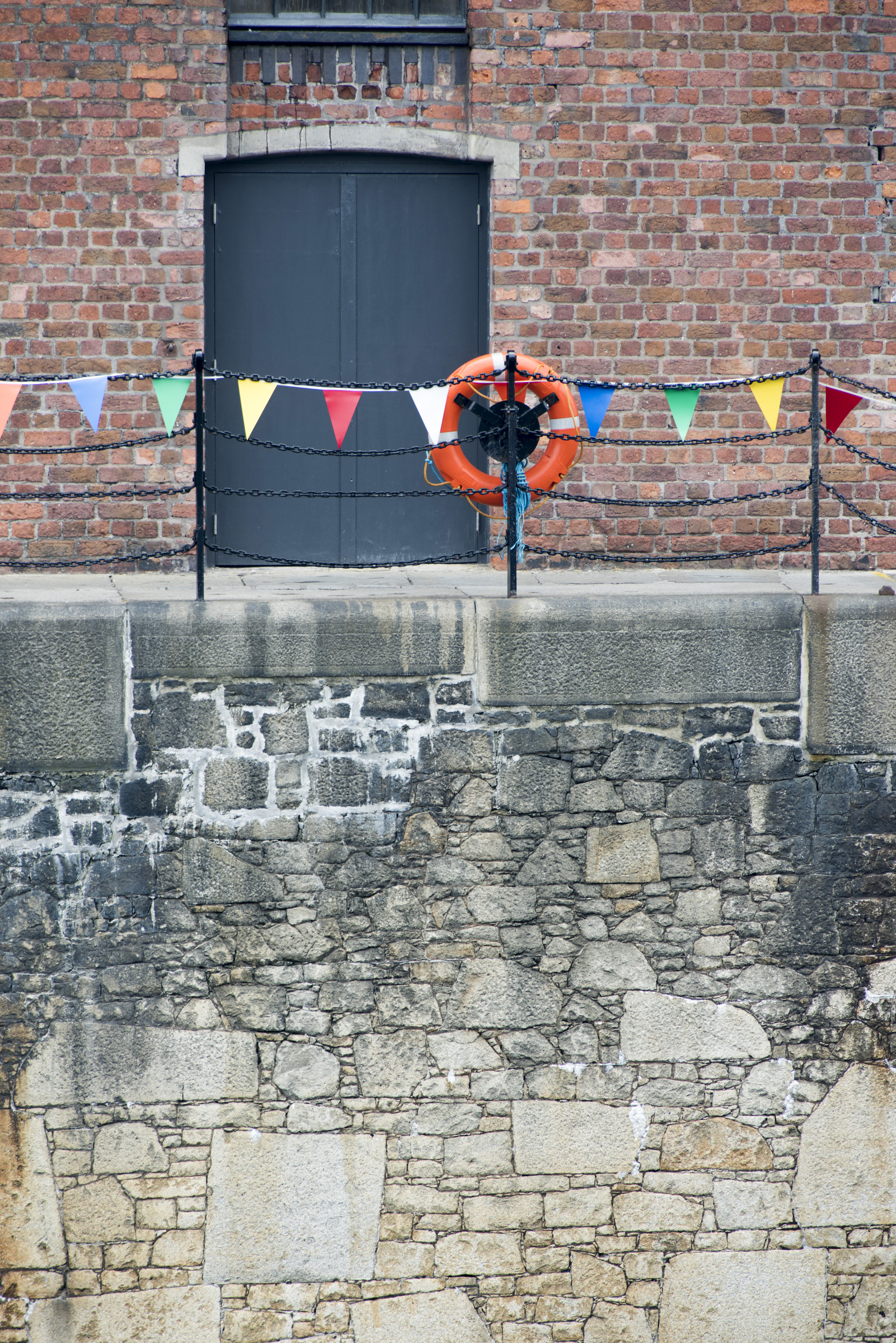 an image of Orange life preserver on iron pole and chain railing at Liverpool Albert Dock with locked black doors surround by brick wall