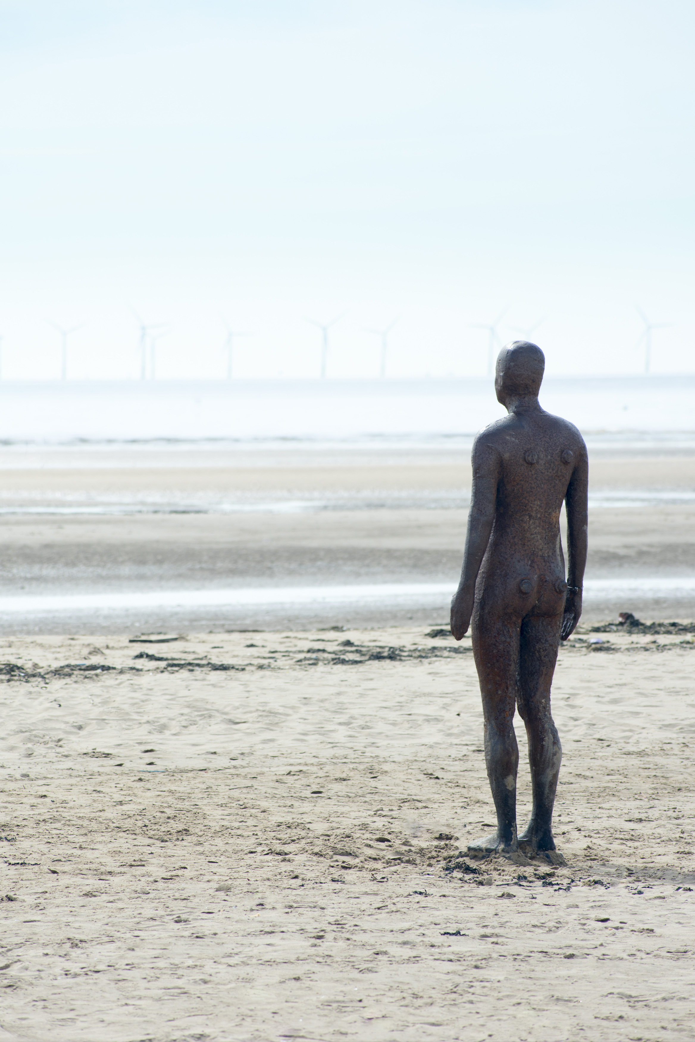 an image of Another Place statue facing empty beach in Crosby, United Kingdom, with windmill power turbines in the far distance