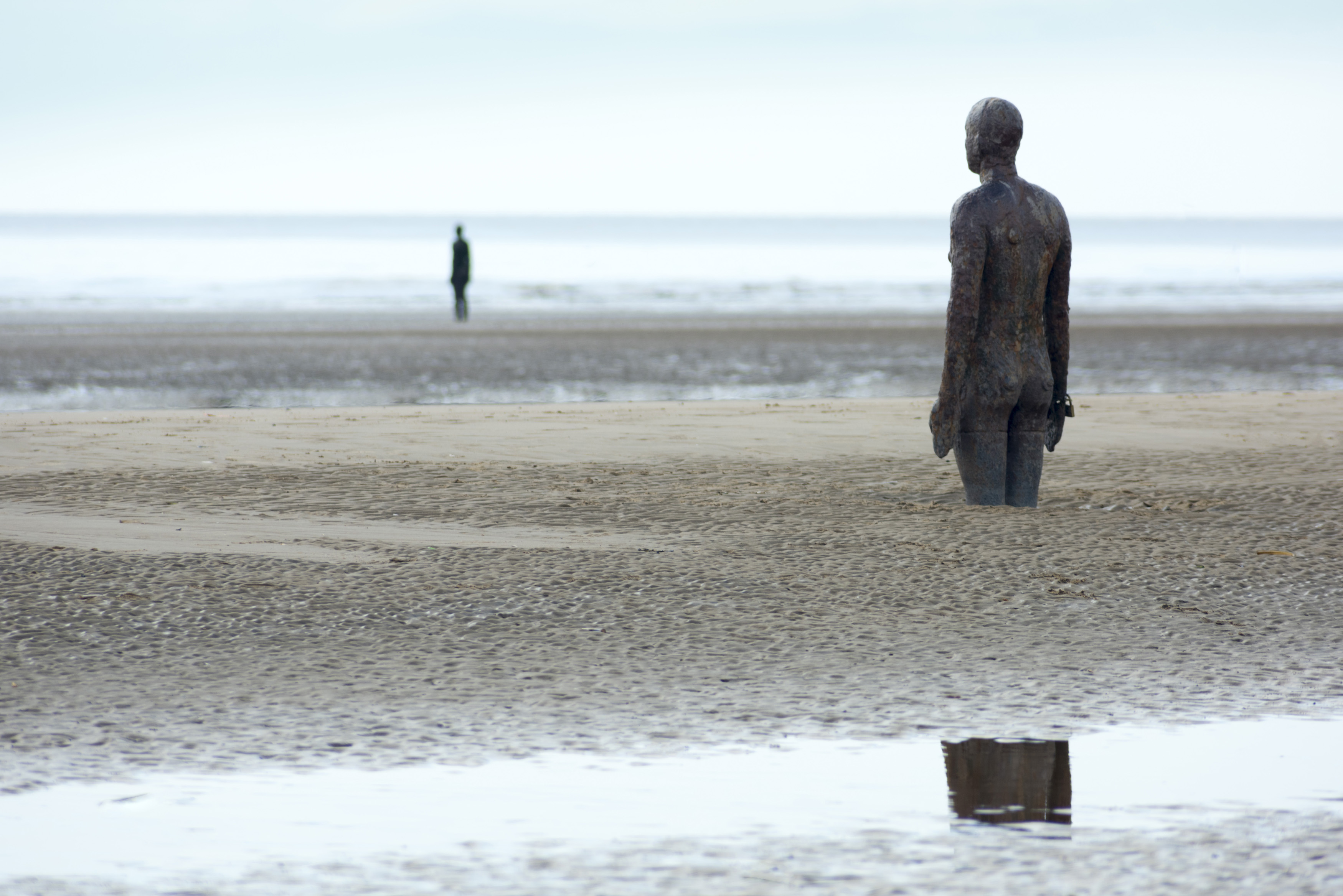 an image of Pair of human shaped standing sculptures partially covered in sand at Crosby, UK under overcast sky