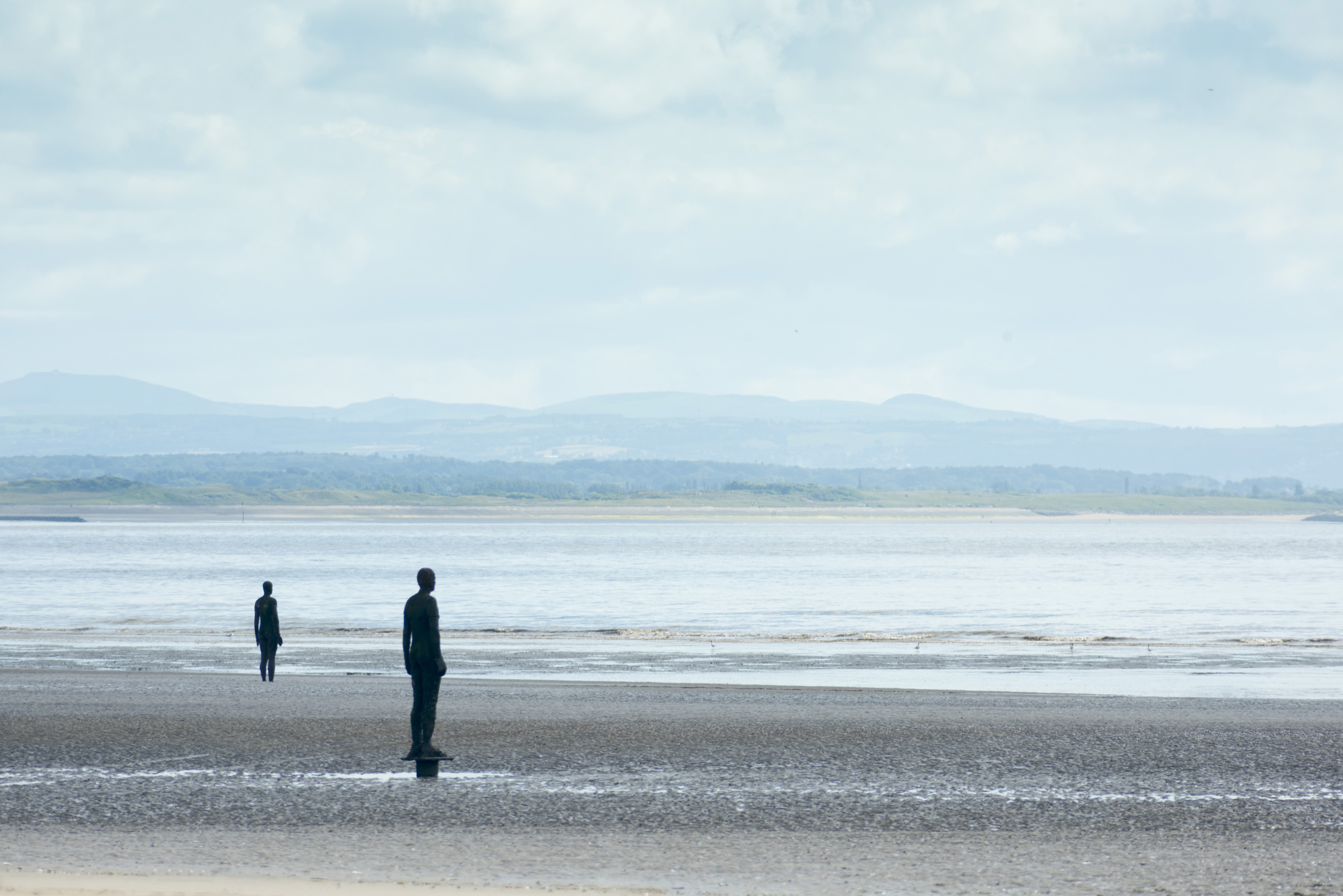 an image of Pair of solitary life size Another Place statues made by Antony Gormley facing water in Crosby, United Kingdom