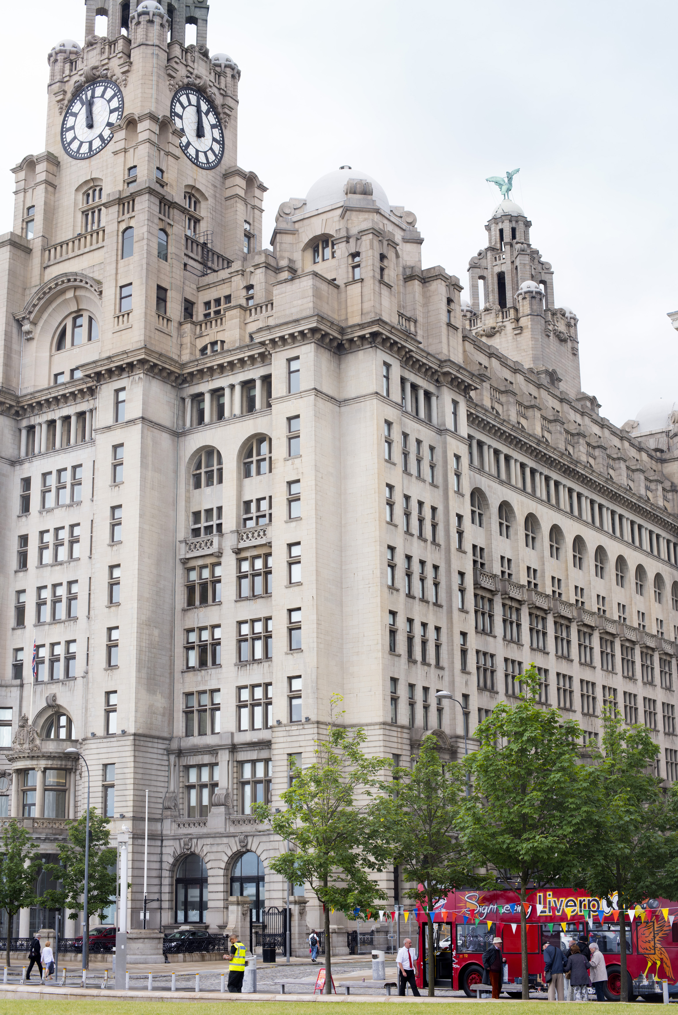 an image of Liverpool Liver Building, a famous landmark in the city and part of the UNESCO designated World Heritage Maritime Mercantile City, with a traditional red tourist bus in the foreground with tourists