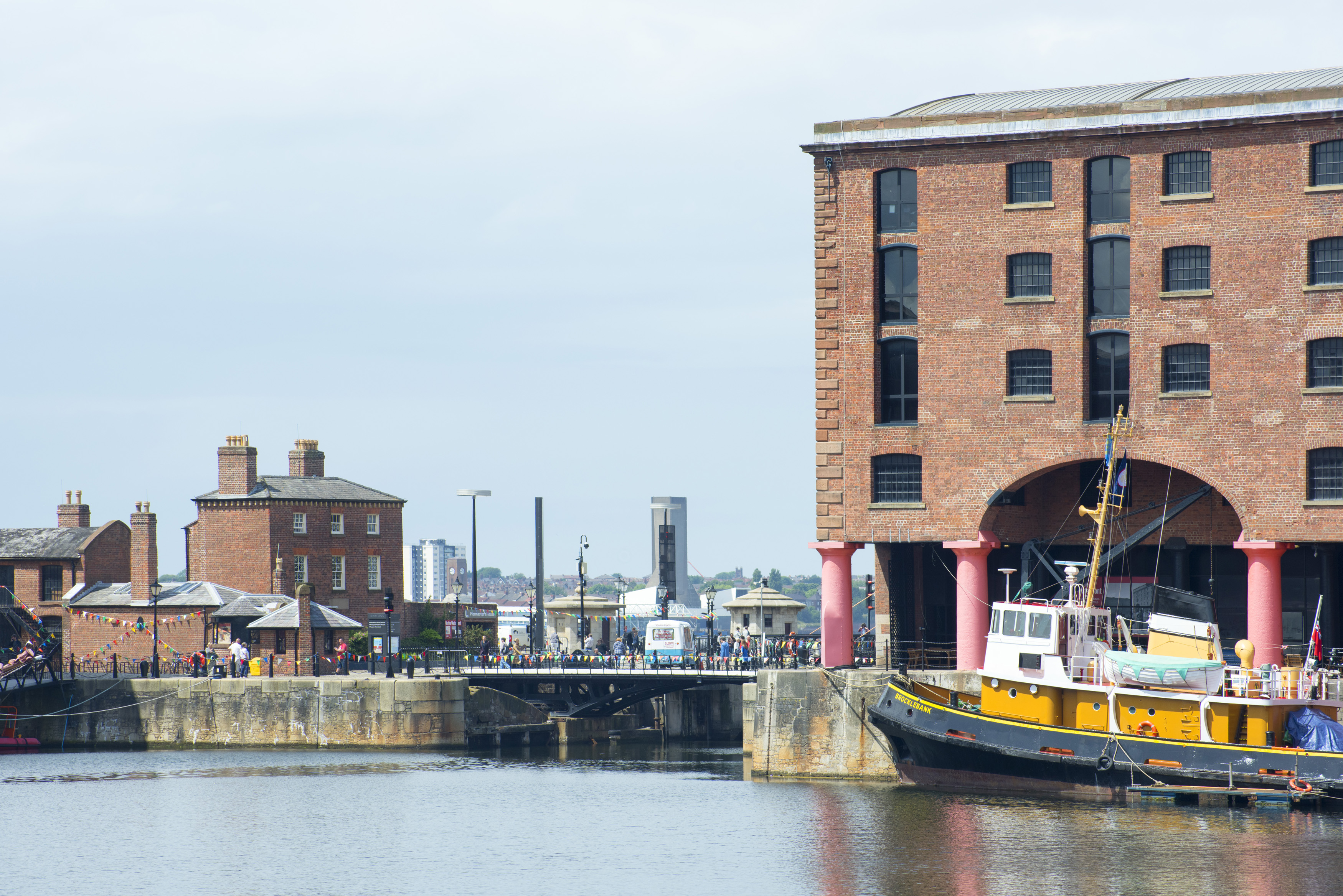 an image of Waterfront view of a quay at Albert Dock, Liverpool, England with an old boat moored alongside, pedestrians and the city skyline behind