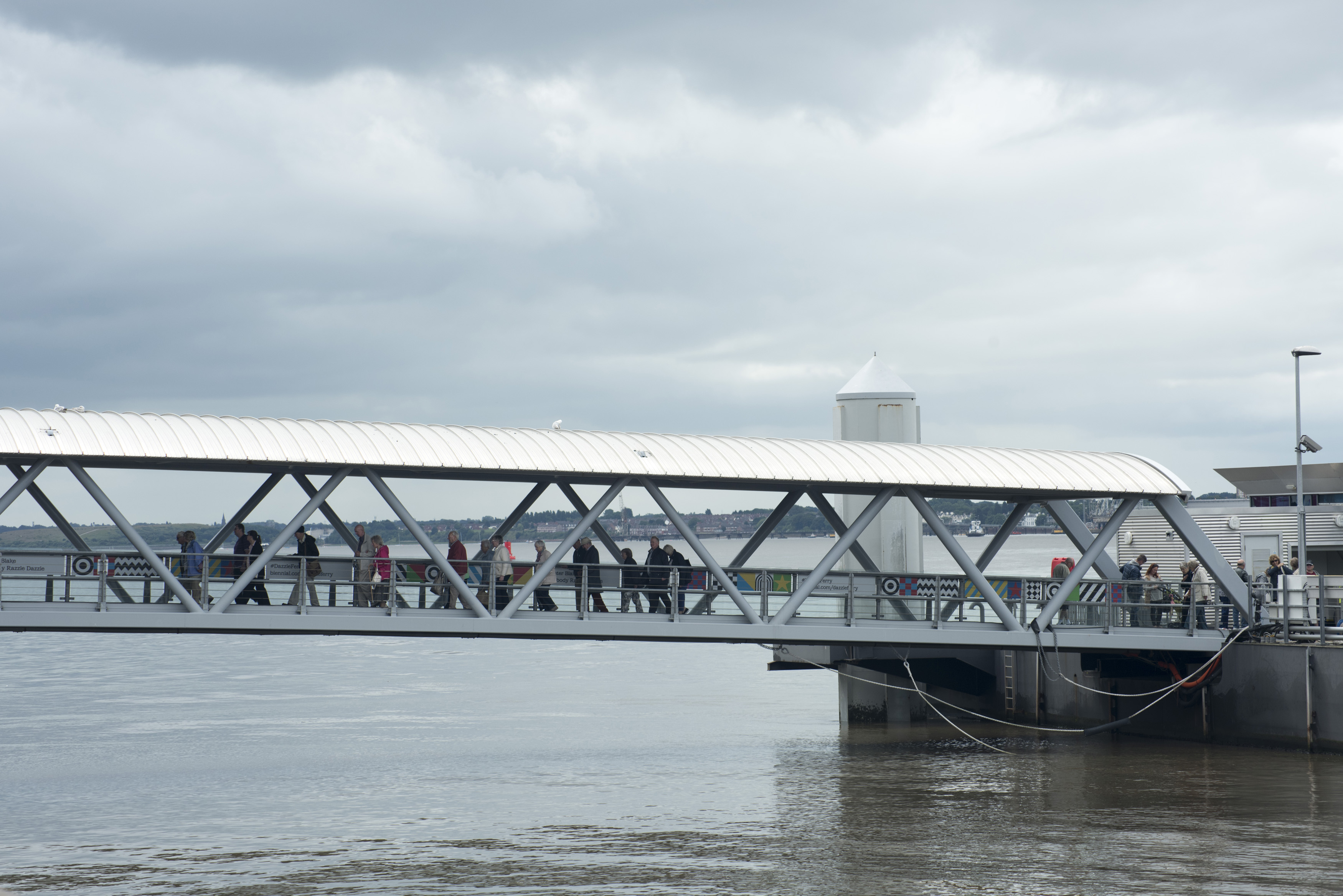 an image of Various unidentifiable tourists traveling across a covered pedestrian walkway bridge over water in Liverpool