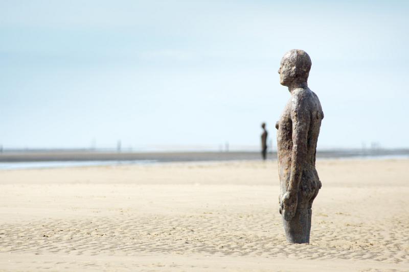 Cast iron statue in the sand on a beach facing out to sea - Another Place, by Antony Gormley at Crosby Beach, Liverpool UK