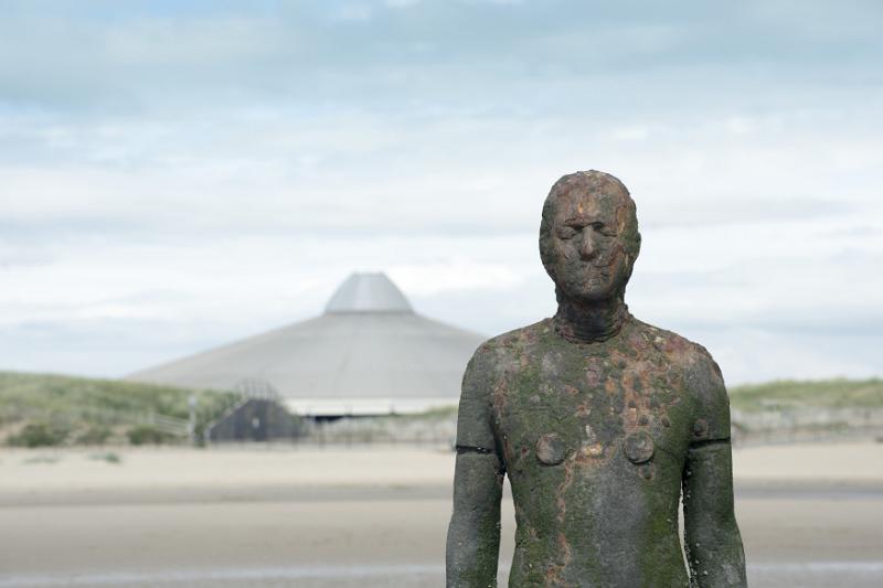 weathered Another Place statue in Crosby, United Kingdom near sandy beach and flat domed building