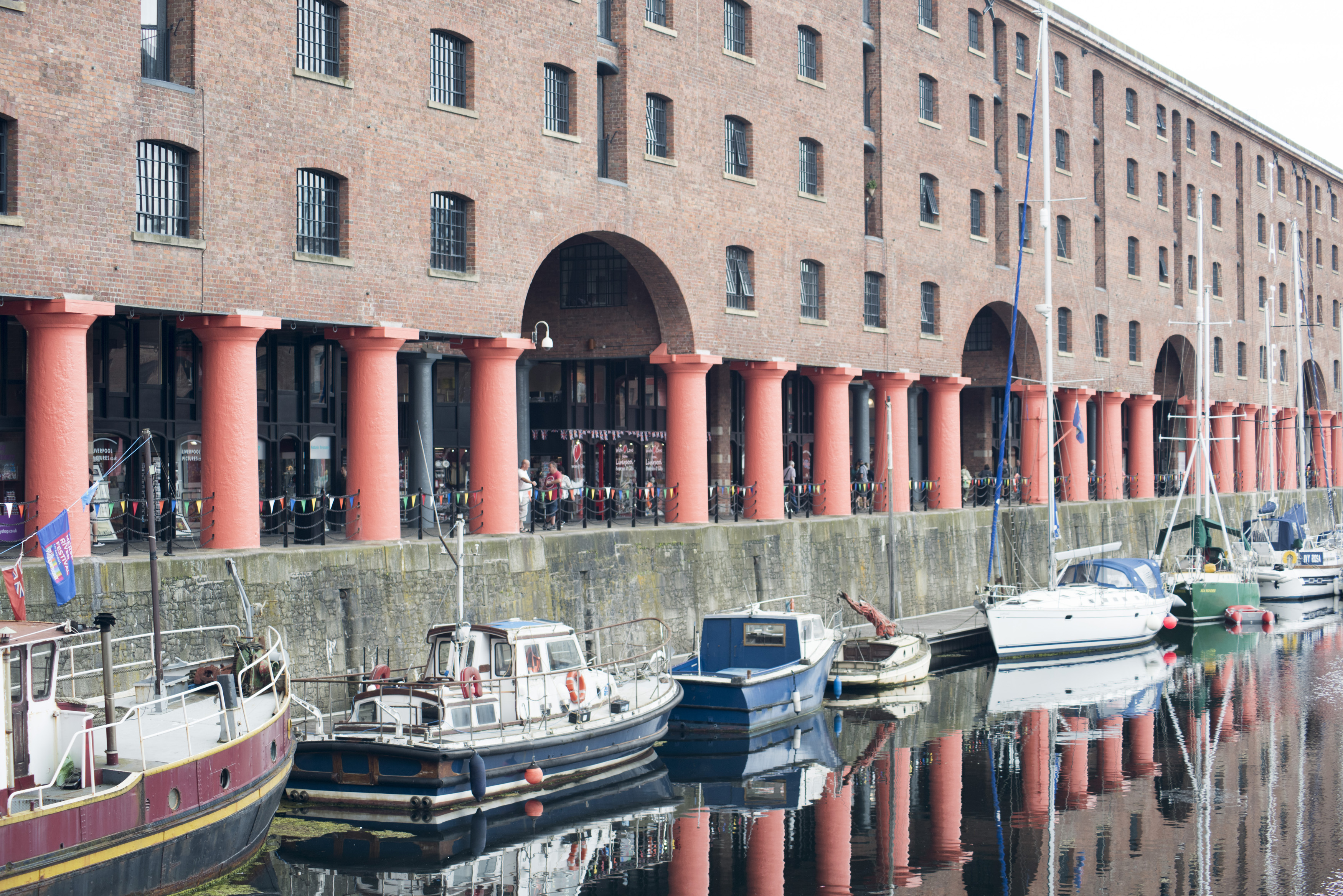 an image of Large red support columns reflecting in water with various little boats docked at Liverpool Albert