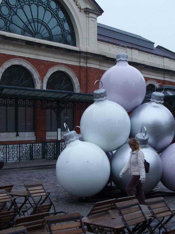 Covent Gardens display of Christmas decorations with a woman walking past a stack of large silver baubles near on open-air restaurant , London, England