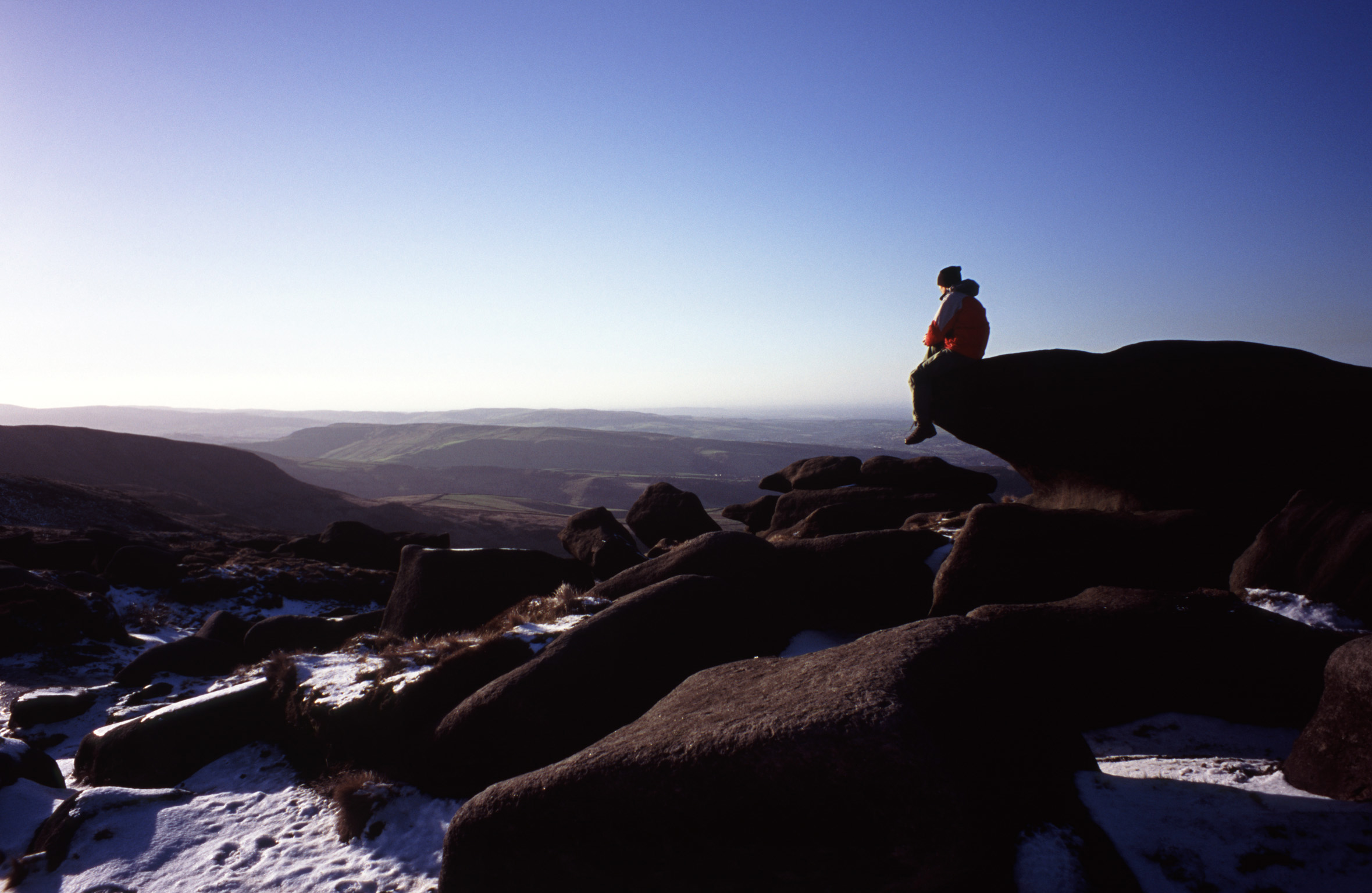 an image of Person enjoying the tranquility of nature sitting on a rock on a snowy winter Kinder Scout, a moorland plateau and National Nature Reserve in the Dark Peak of the Derbyshire Peak District in England