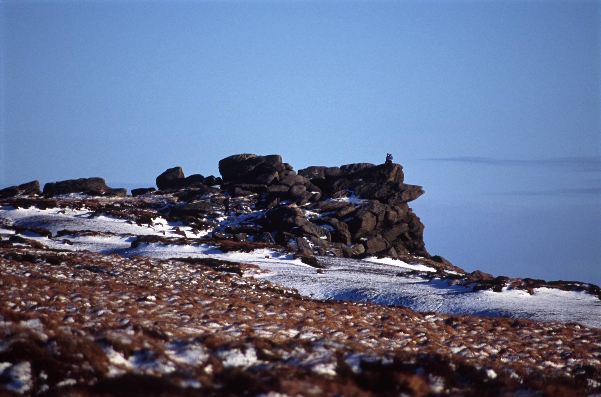 an image of Cold snowy winter walk on Kinder Scout, a moorland plateau and National Nature Reserve in the Dark Peak of the Derbyshire Peak District in England with a rocky outcrop against a clear blue sky