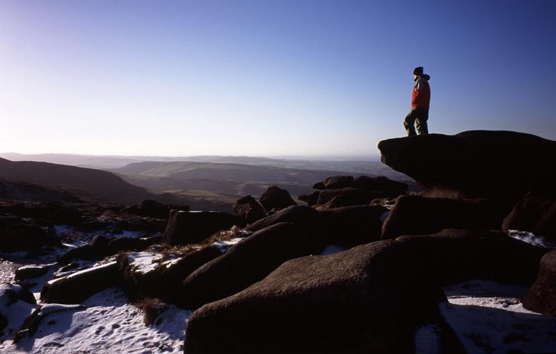 Man hiking on the Kinder Scout Plateau in winter standing on a rocky outcrop admiring the view over the moorlands in the Derbyshire Peak District