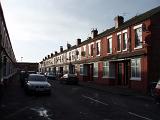 Terraced red brick houses lining a narrow urban street with parked cars in Moss Side , Manchester
