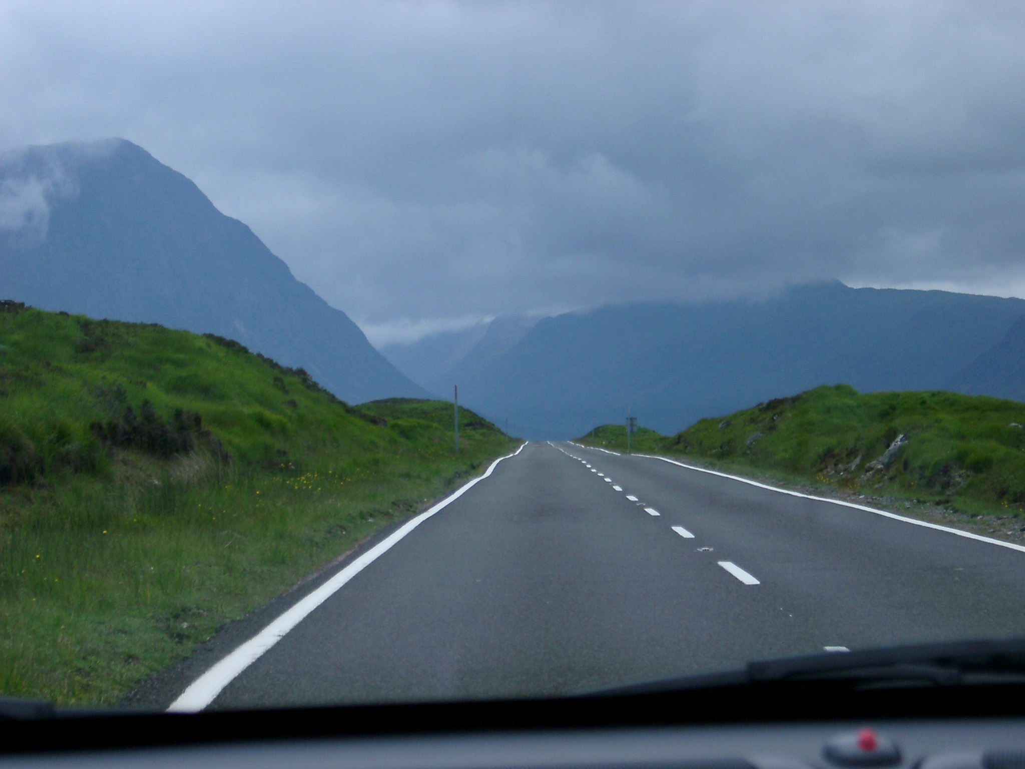 an image of View through the windshield of a car of a tarred road through the Scottish highlands with high mountain peaks on a cold overcast day