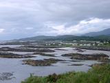 View across coastal grass and stones dotted in shallow water of houses and cottages along the shores of a loch against a moutain background and cloudy grey sky