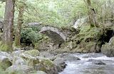 Low angle view over the rocks and flowing water of an arched stone bridge over a river in a leafy green woodland setting