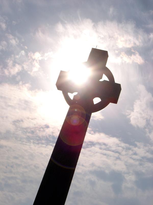 Low angle view looking up at a stone Celtic cross with sun flare silhouetted against a cloudy blue sky