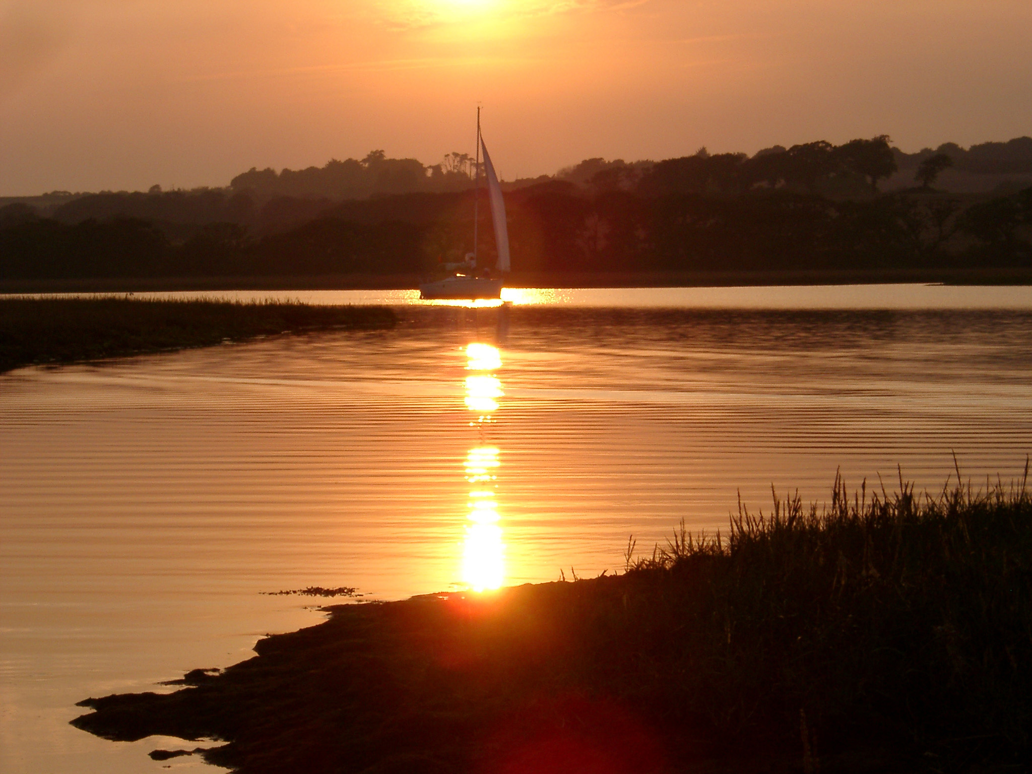 an image of Yacht sailing on tranquil water at sunset passing through a bright orange beam of light reflected on the water