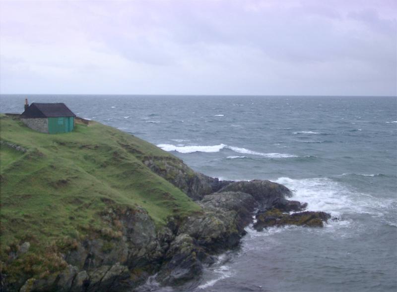 Rocky headland with the roof of a seafront cottage just visible jutting into a calm ocean on an overcast cloudy day