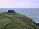 Stormy coastal landscape with the roof of a small hut or cottage on a lush promontory with pastures overlooking a wild ocean with breaking waves