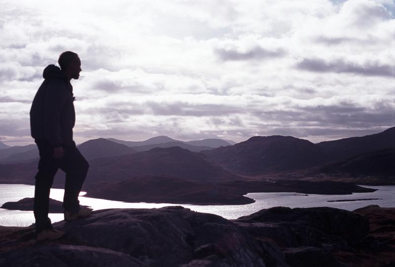 Climber silhouetted on a mountain peak overlooking a scenic landscape with lakes at dusk against a cloudy sky