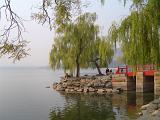 People standing under green weeping willow trees on a red wooden bridge to a stone headland on a tranquil scenic lake