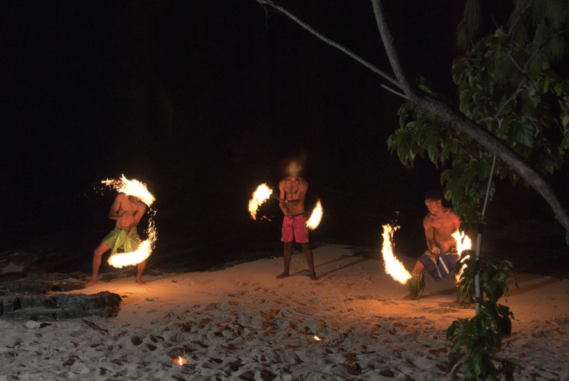 an image of Three Fijian men performing a fire dance on the beach twirling flaming branches in the night sky