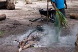 Man tending to a lovo, or earth oven, which is a cooking pit in the ground which is covered with hot coals and charcoal which he is fanning with a palm frond