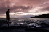 Man in silhouette standing on the seashore watching a marine sunset with clouds tinted a lilac purple