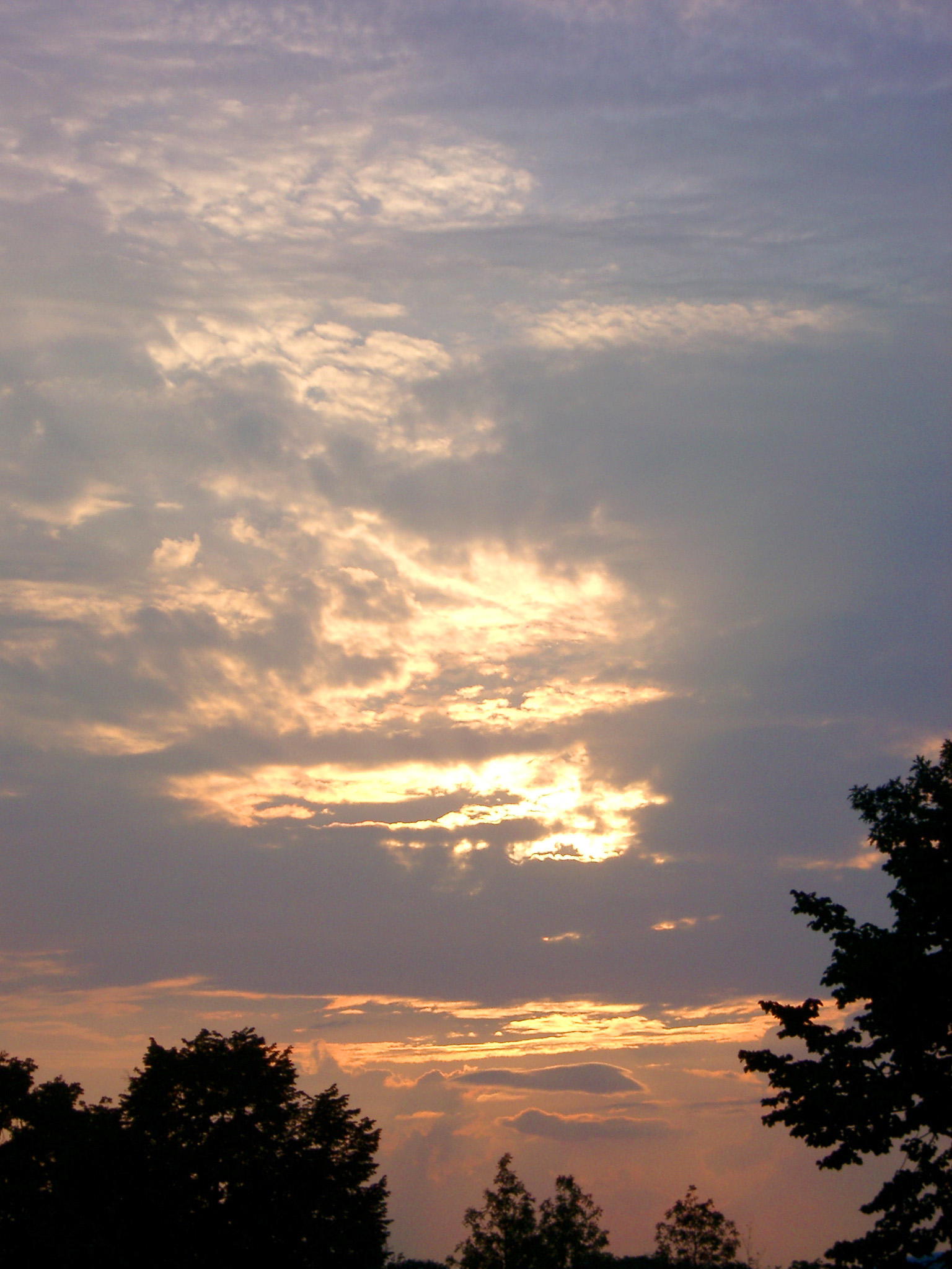 an image of Fiery sunset in an overcast sky with the sun peeking through the layers of cloud over evergreen trees