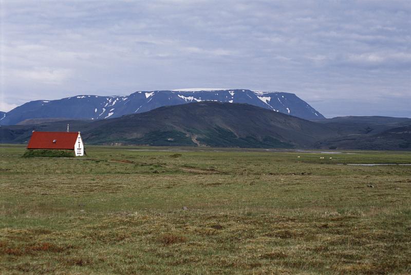 Remote red roofed hut in Iceland standing in open grassland with distant snow covered mountain peaks