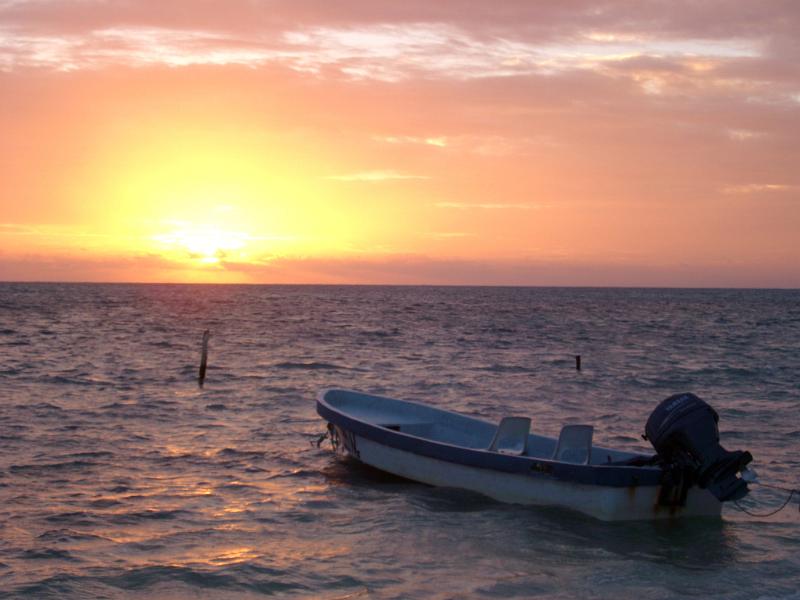 Colorful Caribbean sunset with a glowing orange sun dipping below the ocean and a moored wooden motorboat in the foreground