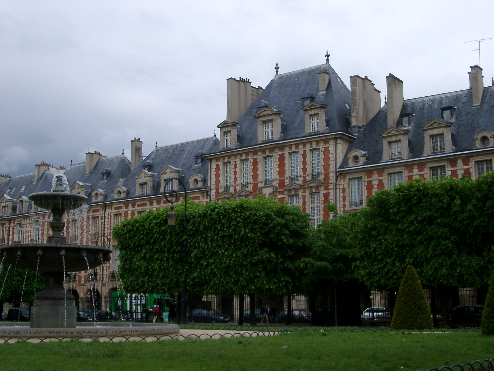 an image of Green Landscape with Trees and Water Fountain Fronting Famous Vintage Place de Vosges Buildings. Isolated on Lighter Blue Gray Sky Background.