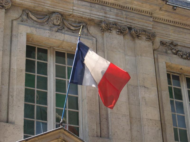The Tricolor French National Flag in red, white and blue flying from a flagpole on the exterior of an old stone building