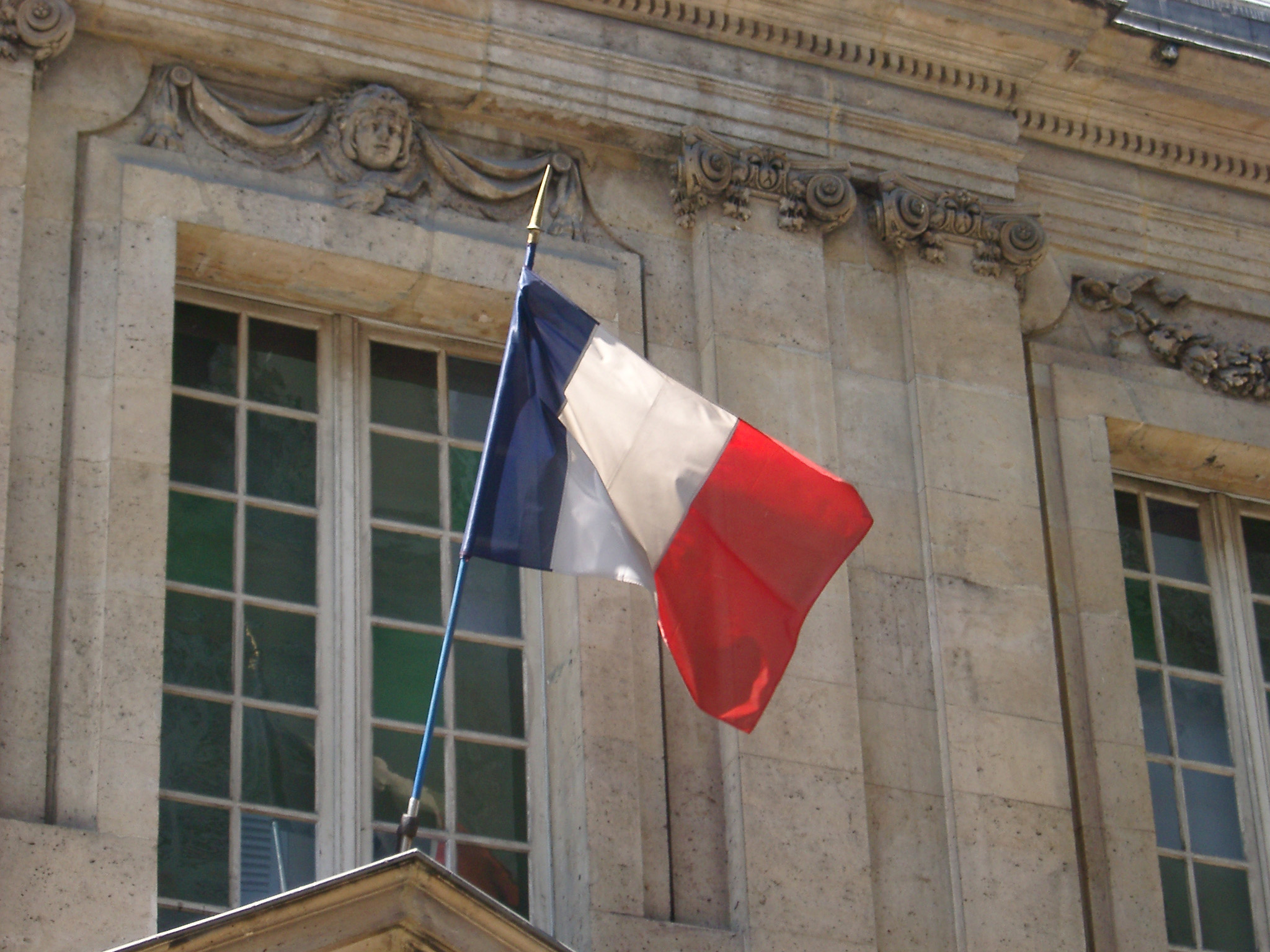 an image of The Tricolor French National Flag in red, white and blue flying from a flagpole on the exterior of an old stone building