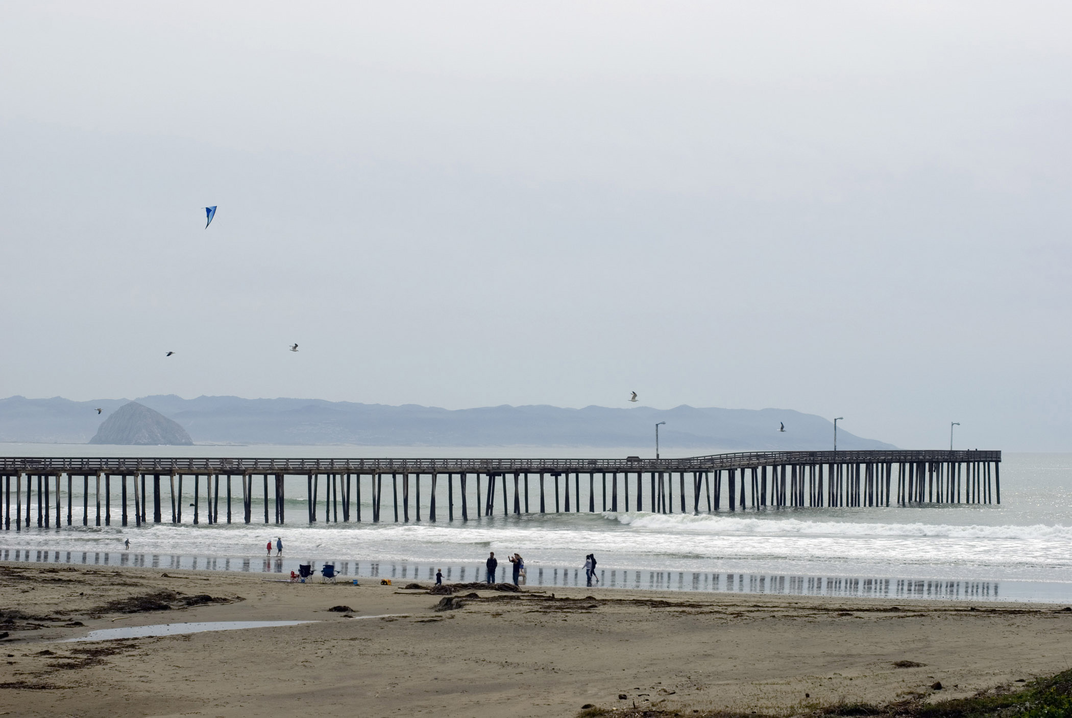 an image of Tourists at Beautiful Cayucos Pier. Captured in Extensive View with Morro Rock Afar. Isolated on Foggy Sky Background.