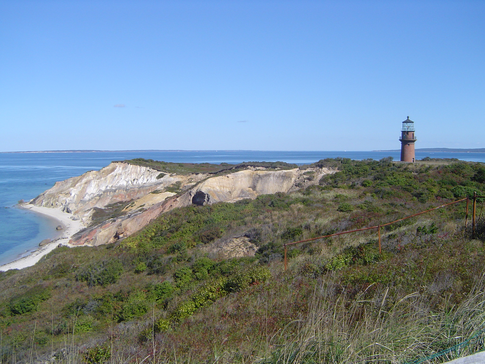 an image of Grassy Ocean Cliff with Lighthouse Afar in Extensive View. Isolated on Light Blue Sky Background.
