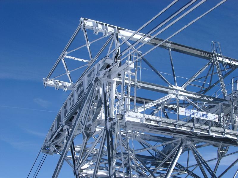 Detail of a cable car tower showing the steel framework and cables on a ski slope against a sunny blue sky
