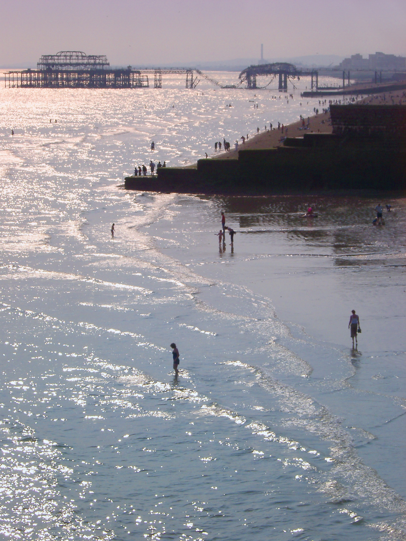 an image of People paddling at Brighton beach with the remnants of the burnt out historical pier visible behind them on a warm summers