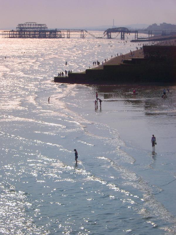 People paddling at Brighton beach with the remnants of the burnt out historical pier visible behind them on a warm summers