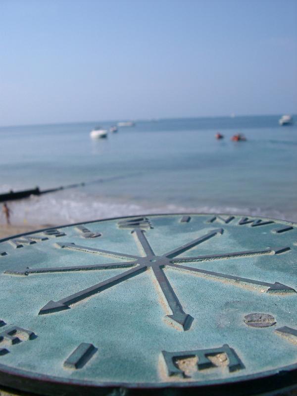 Low angle view over the dial showing the compass points of a brass marine compass overlooking the ocean and moored small boats