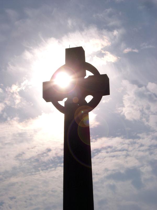Celtic memorial stone cross backlit and silhouetted by the sun with a bright sunflare , low angle against a cloudy blue sky