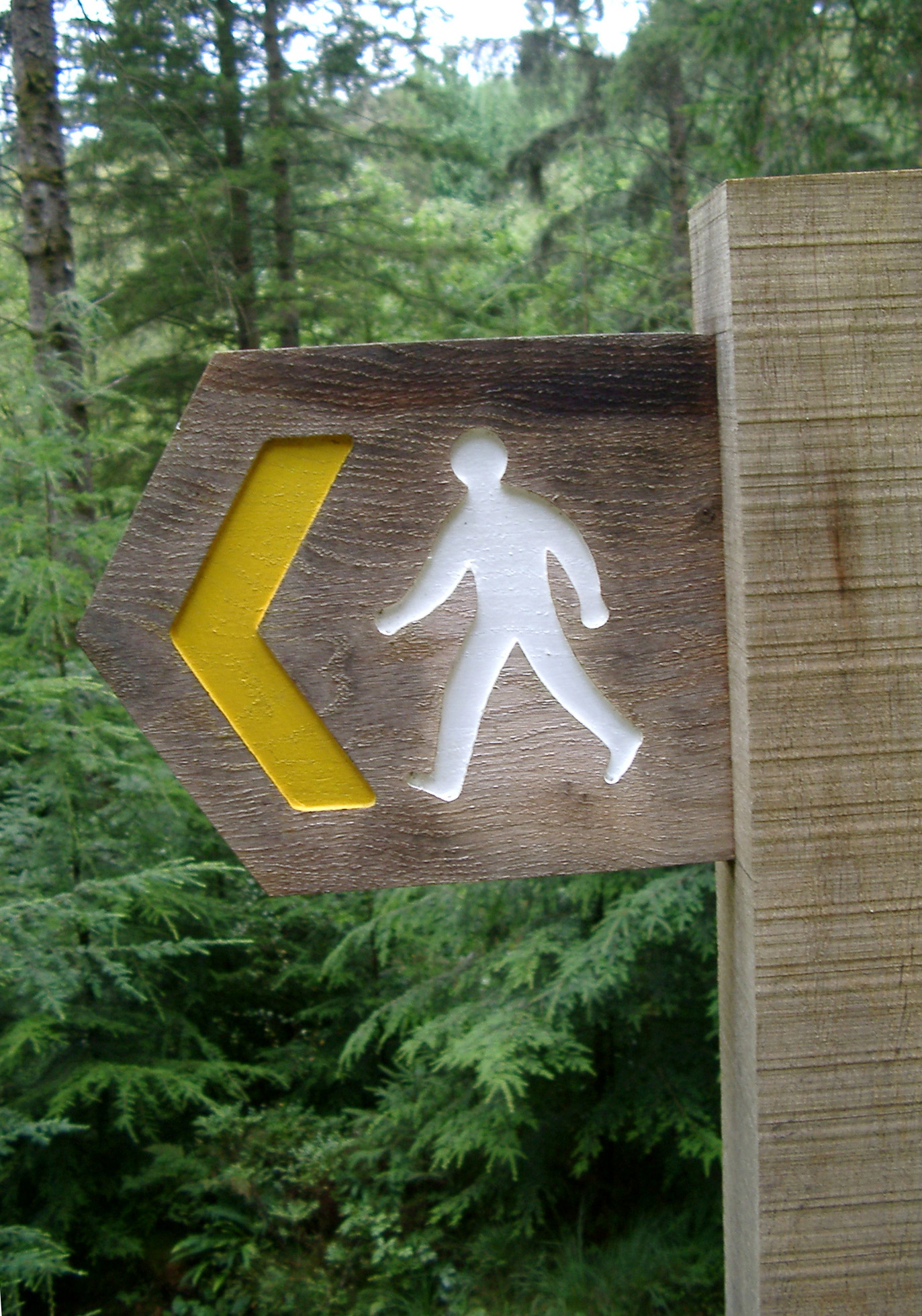 an image of Rustic wooden footpath sign with a silhouette of a walking man and left pointing yellow arrow showing the direction of the trail, outdoors in woodland