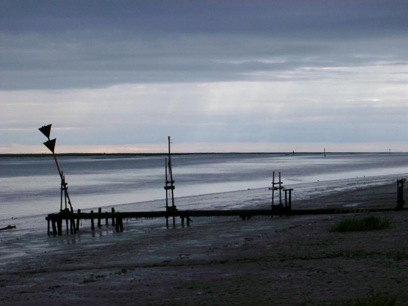 Estuary Dark atmospheric landscape of a marine estuary on an overcast stormy day with a silhouette of a small jetty and warning signs on the beach in the foreground