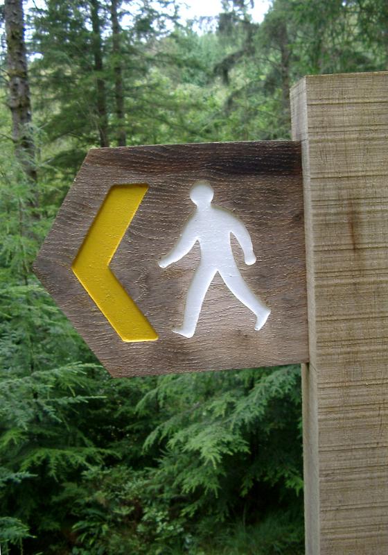 Rustic wooden footpath sign with a silhouette of a walking man and left pointing yellow arrow showing the direction of the trail, outdoors in woodland